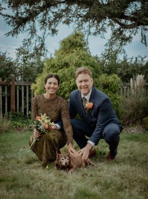 Two people posing with their dog on grassy lawn with trees in background.