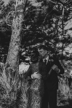 A couple embraces among tall evergreen trees in a moody forest setting.