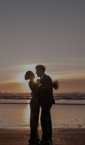 Couple in silhouette share a romantic moment on beach at golden hour with waves crashing behind them.