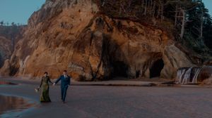 Two figures walk along a scenic beach path with dramatic cliffs and rock formations in the background.