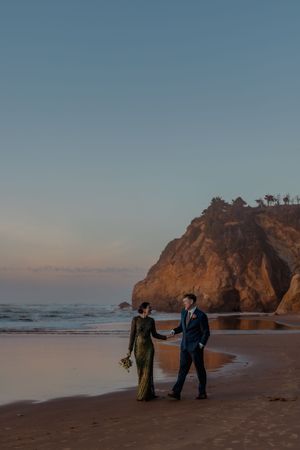 A couple walks hand in hand on a beach at sunset with dramatic cliffs in the background.