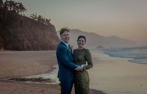 Romantic beach photoshoot with couple embracing near driftwood as sun sets over coastal mountains.
