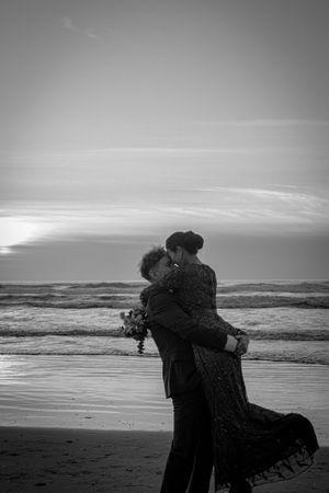 Silhouetted figures embrace on a moody beach at sunset with rolling waves in the background in black and white.