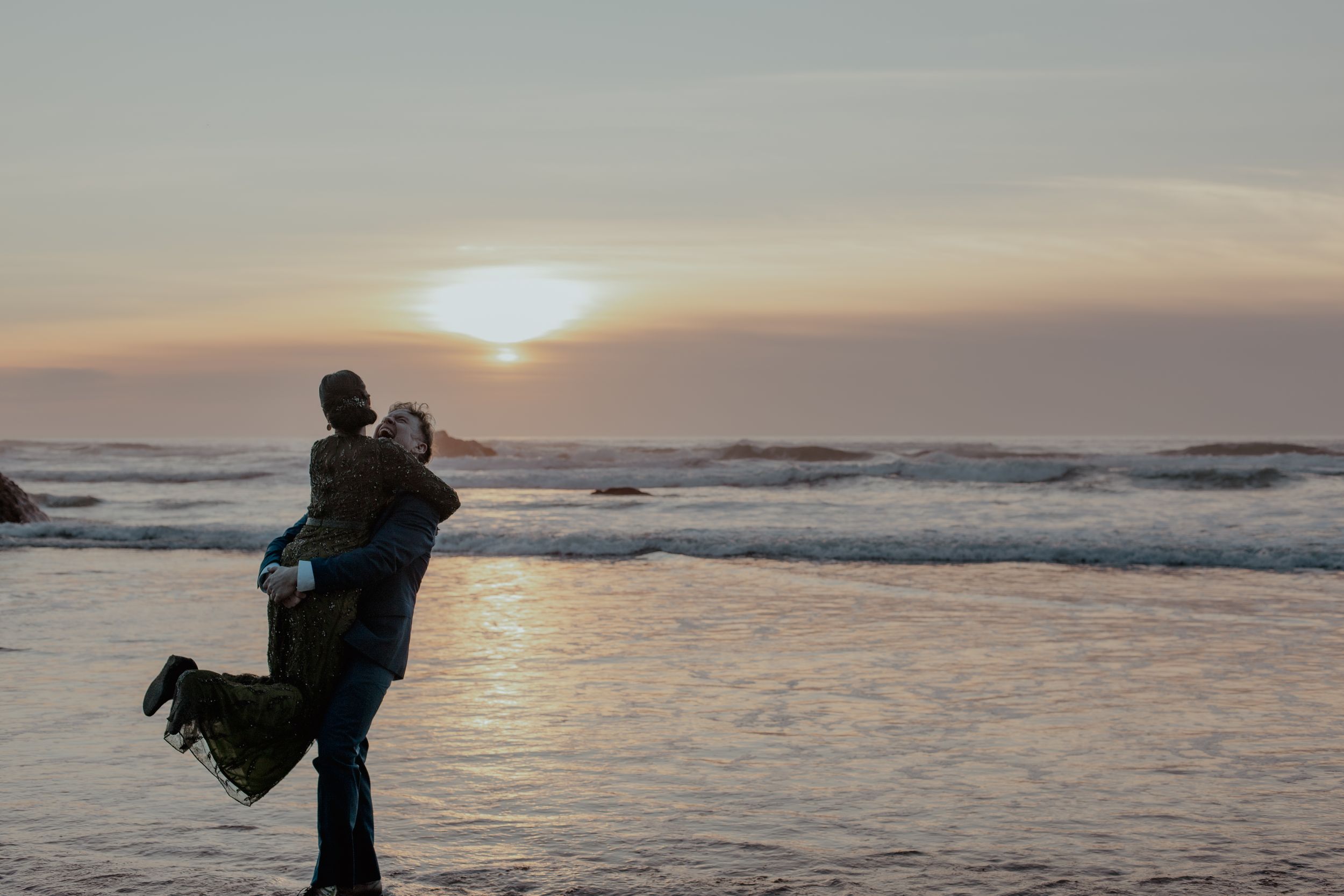 Silhouetted couple embraces on a beach at sunset with ocean waves in the background.