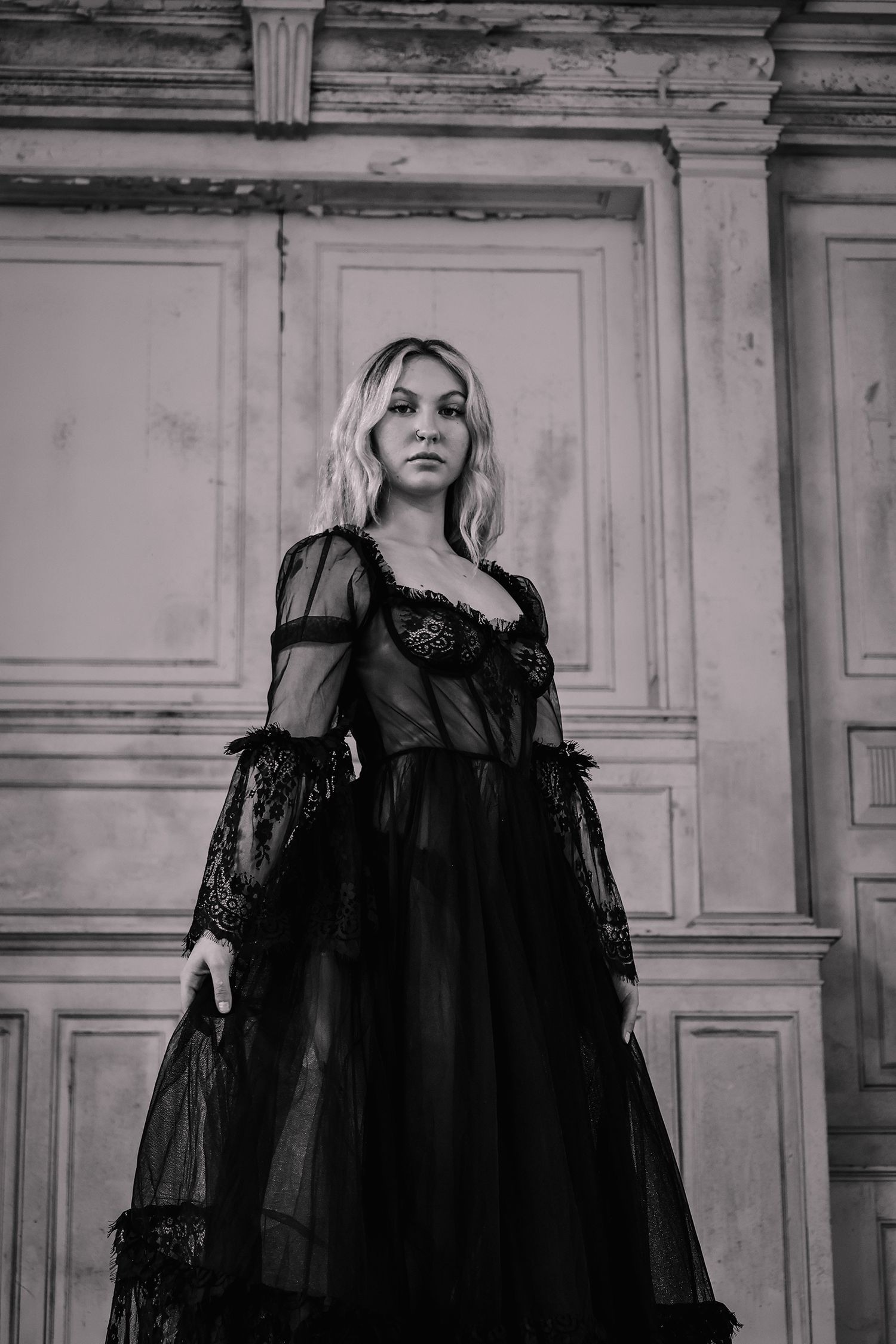 A black and white photo of a witchy woman in a dramatic gothic style black lace gown standing against ornate vintage doors.