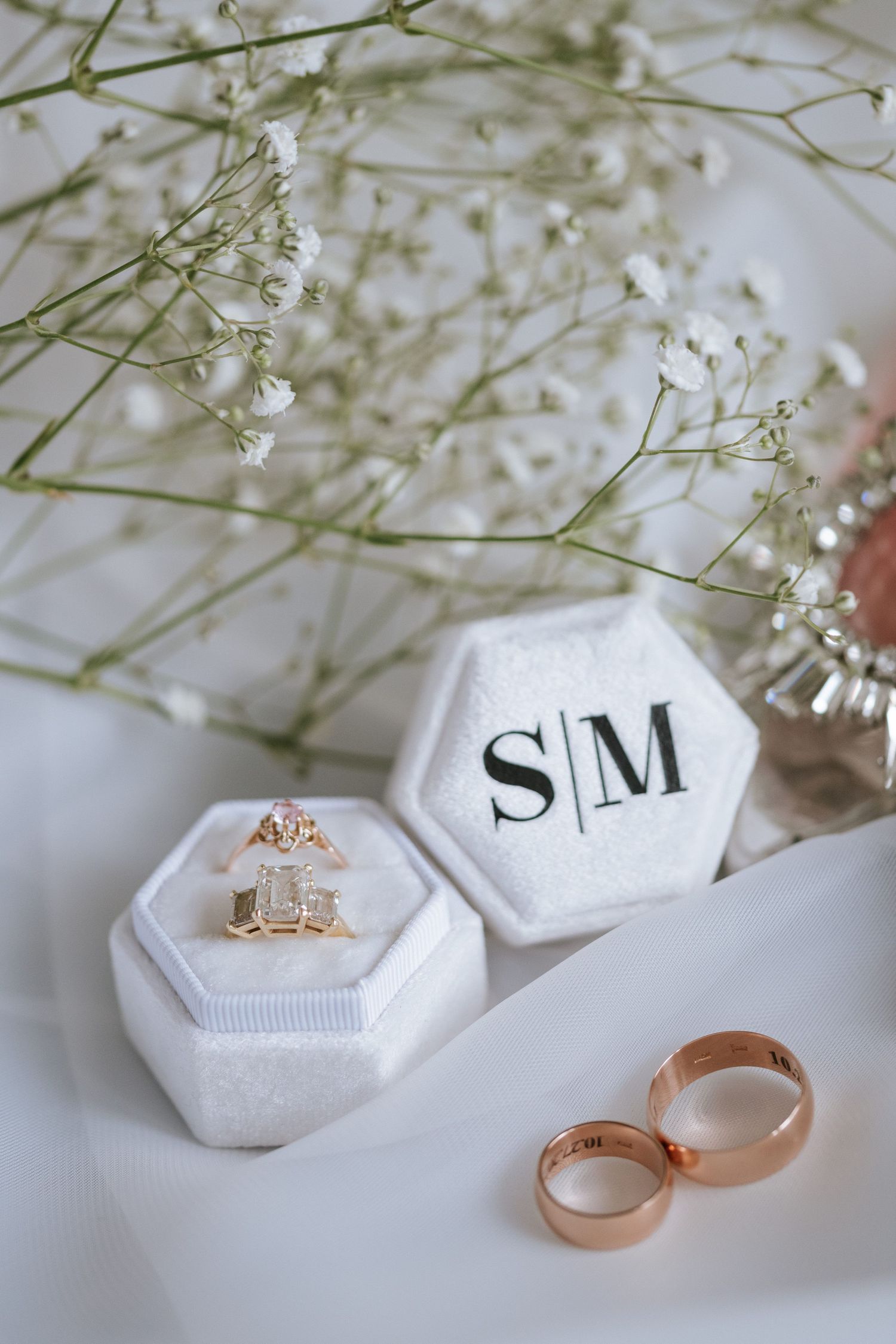 White hexagonal ring boxes with initials and rose gold wedding bands displayed with delicate baby's breath flowers.