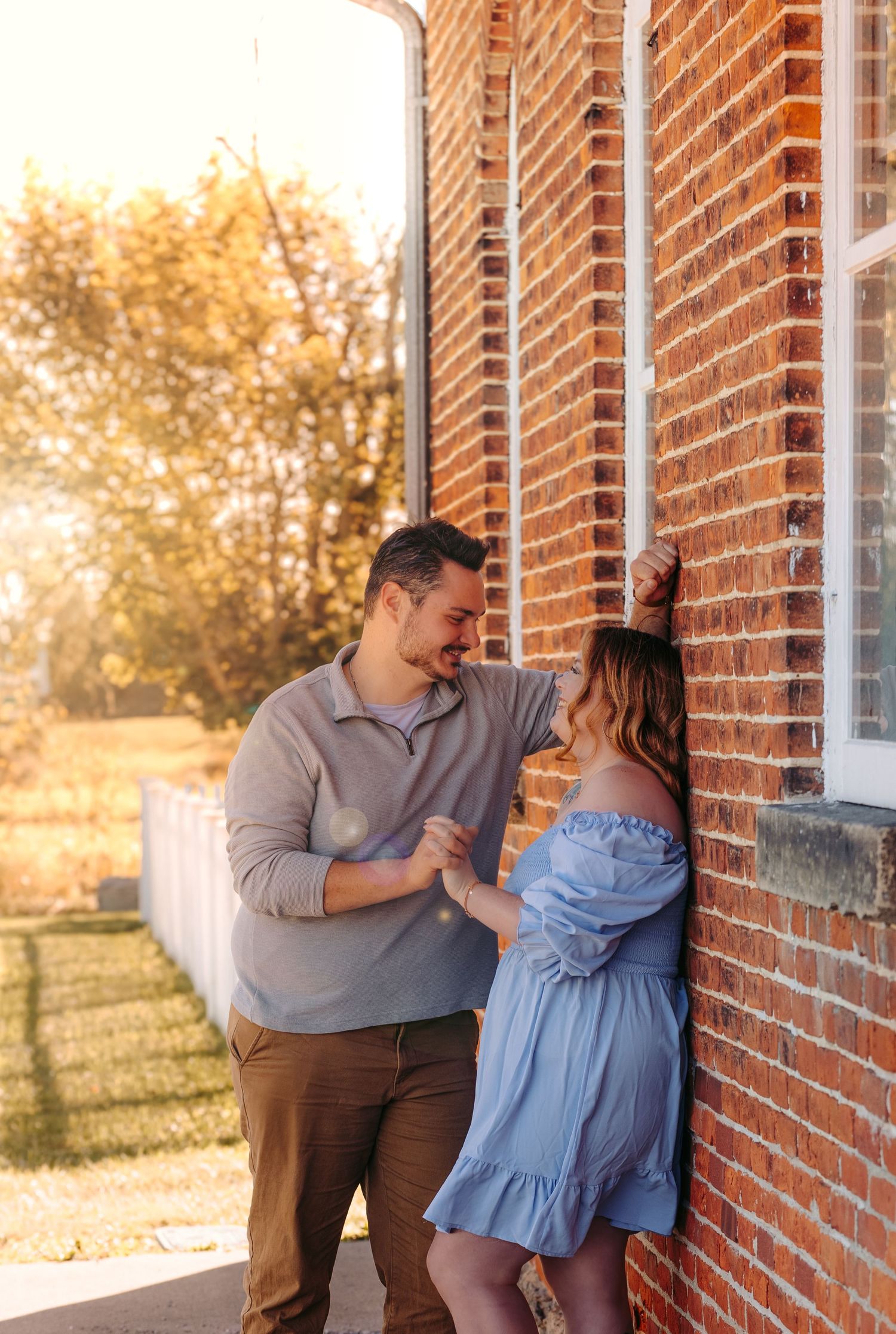 A romantic couple shares an intimate moment against a red brick wall at sunset.