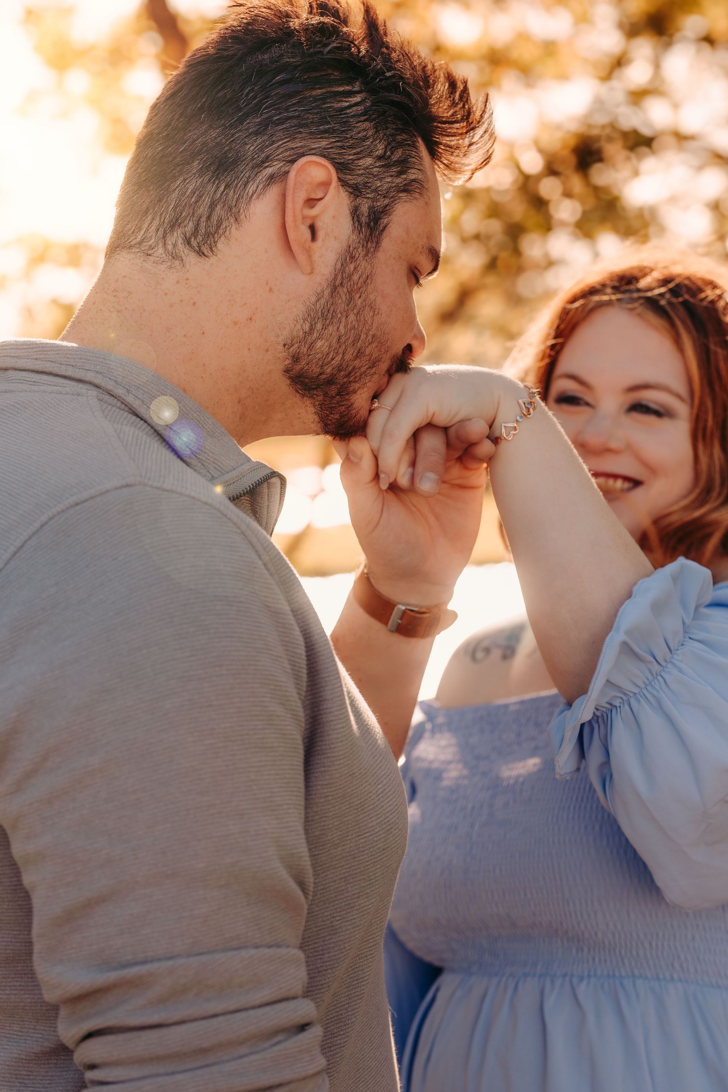 An intimate engagement photo with someone kissing their partner's hand in golden sunlight.