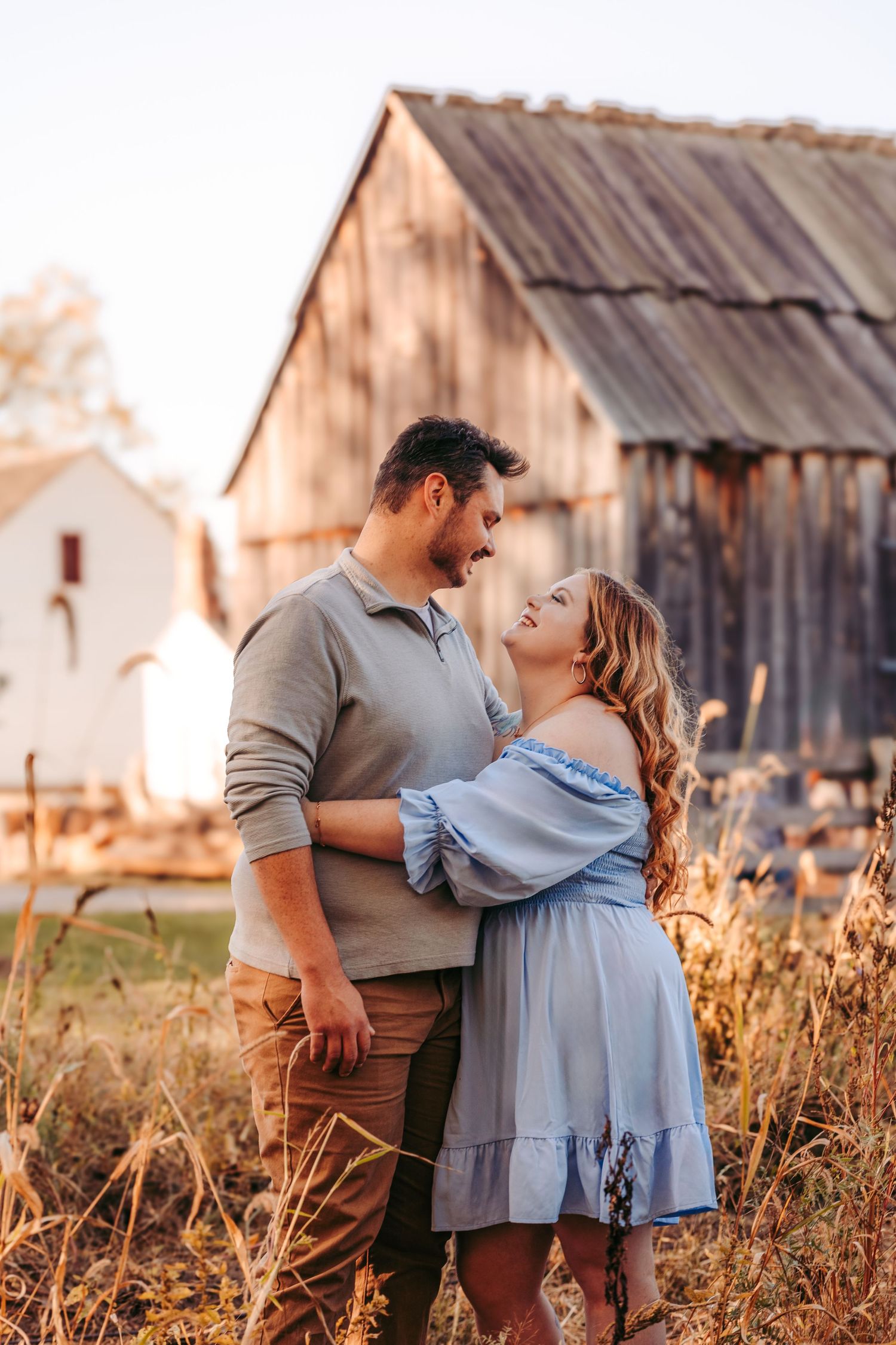 Couple embracing in front of rustic barn during sunset golden hour.