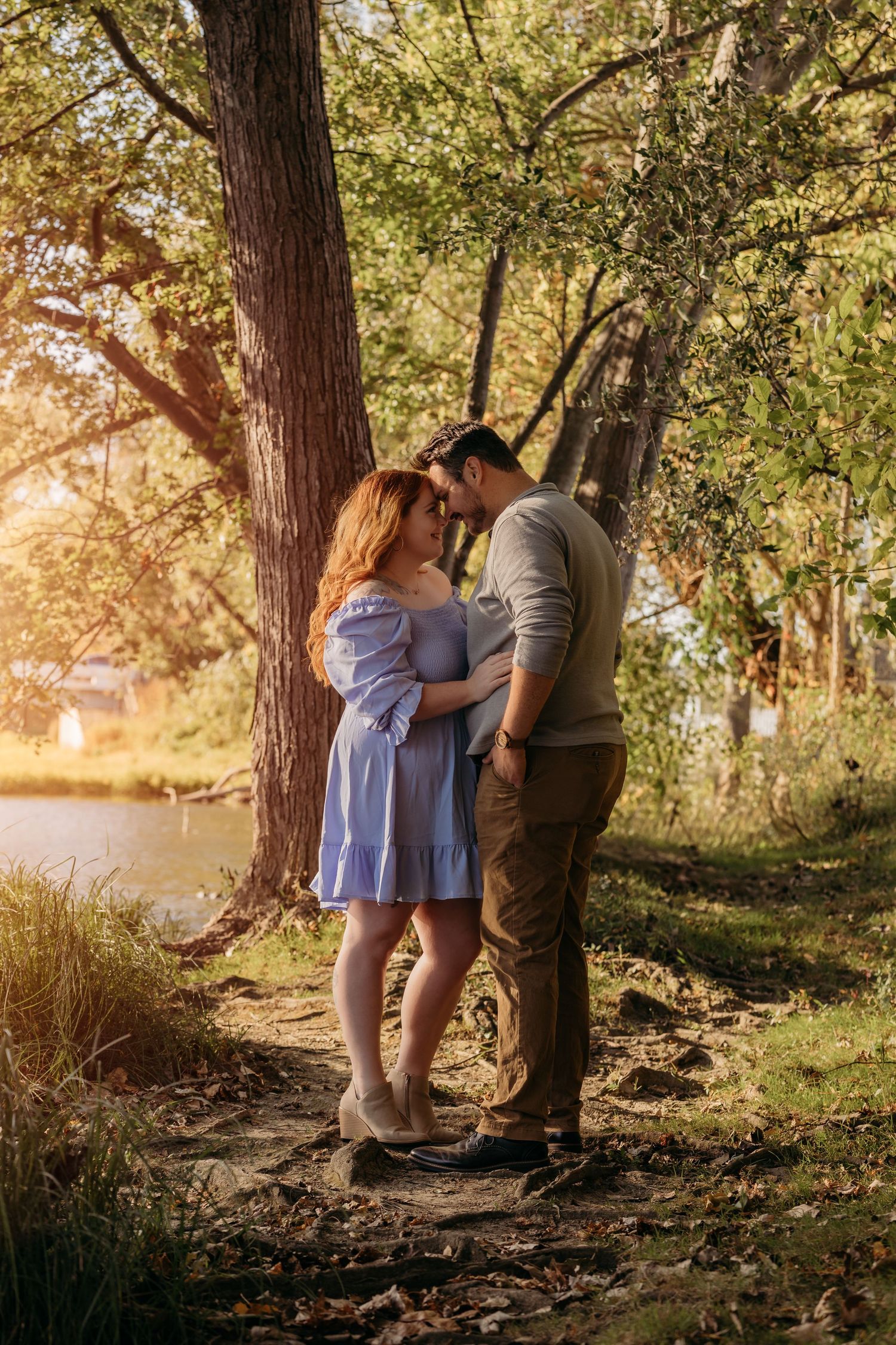 Intimate moment between couple standing by tree near waterfront during golden hour.