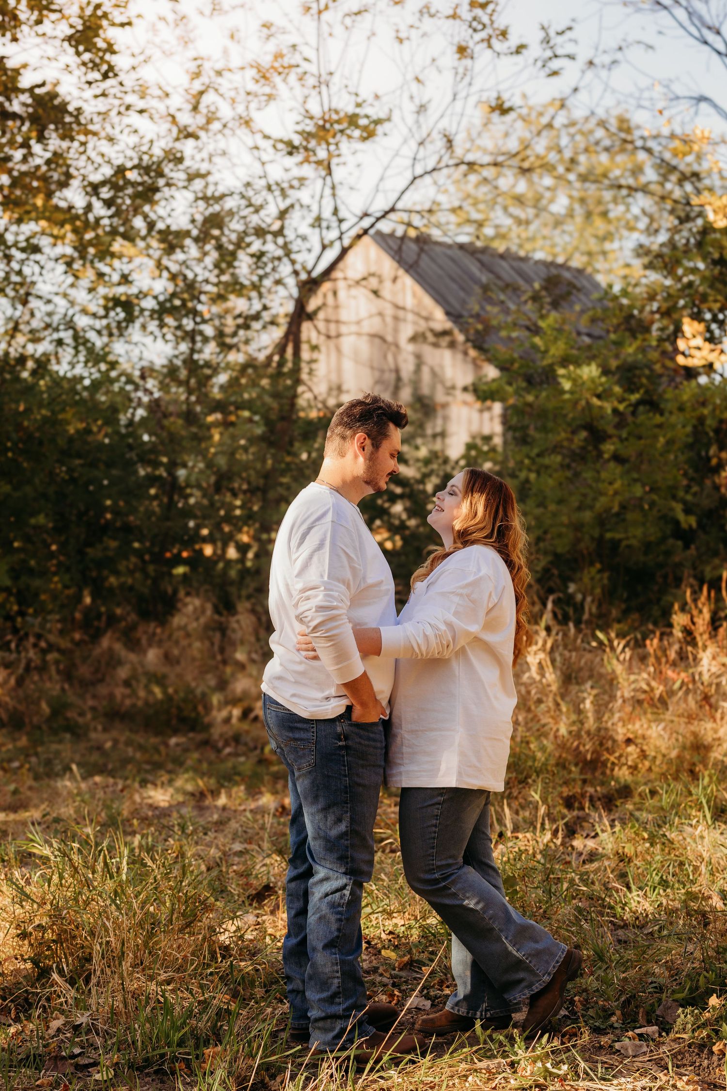Couple standing together in autumn field with old barn in background.