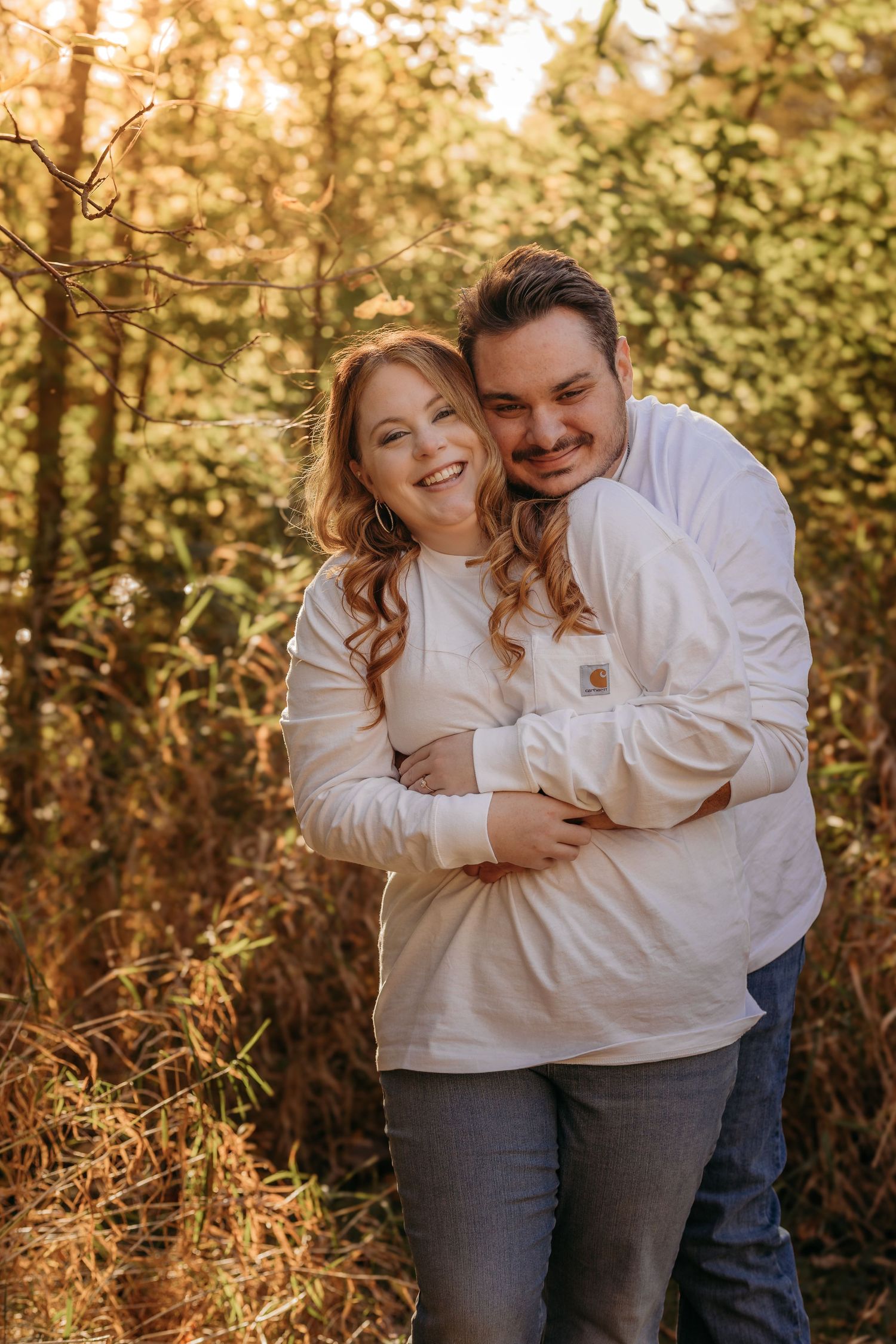 A romantic autumn engagement photo of a couple embracing outdoors in matching white sweaters with golden sunlight.