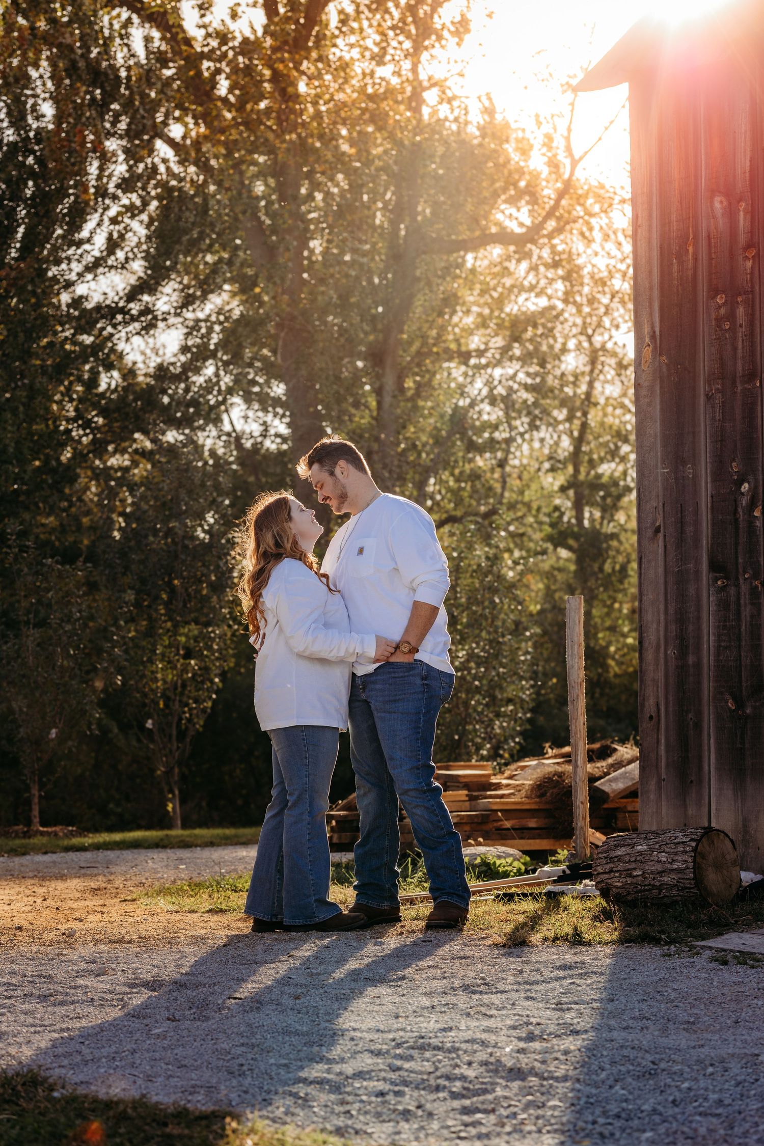 A couple shares an intimate moment at sunset near a rustic barn in casual attire.
