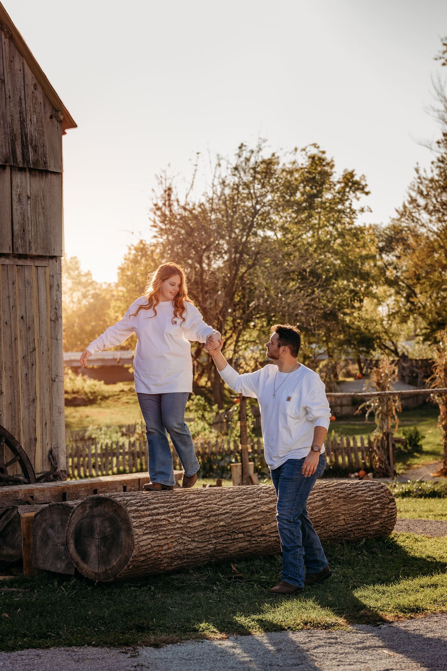 Playful engagement photo with couple holding hands on log near rustic barn in autumn setting.