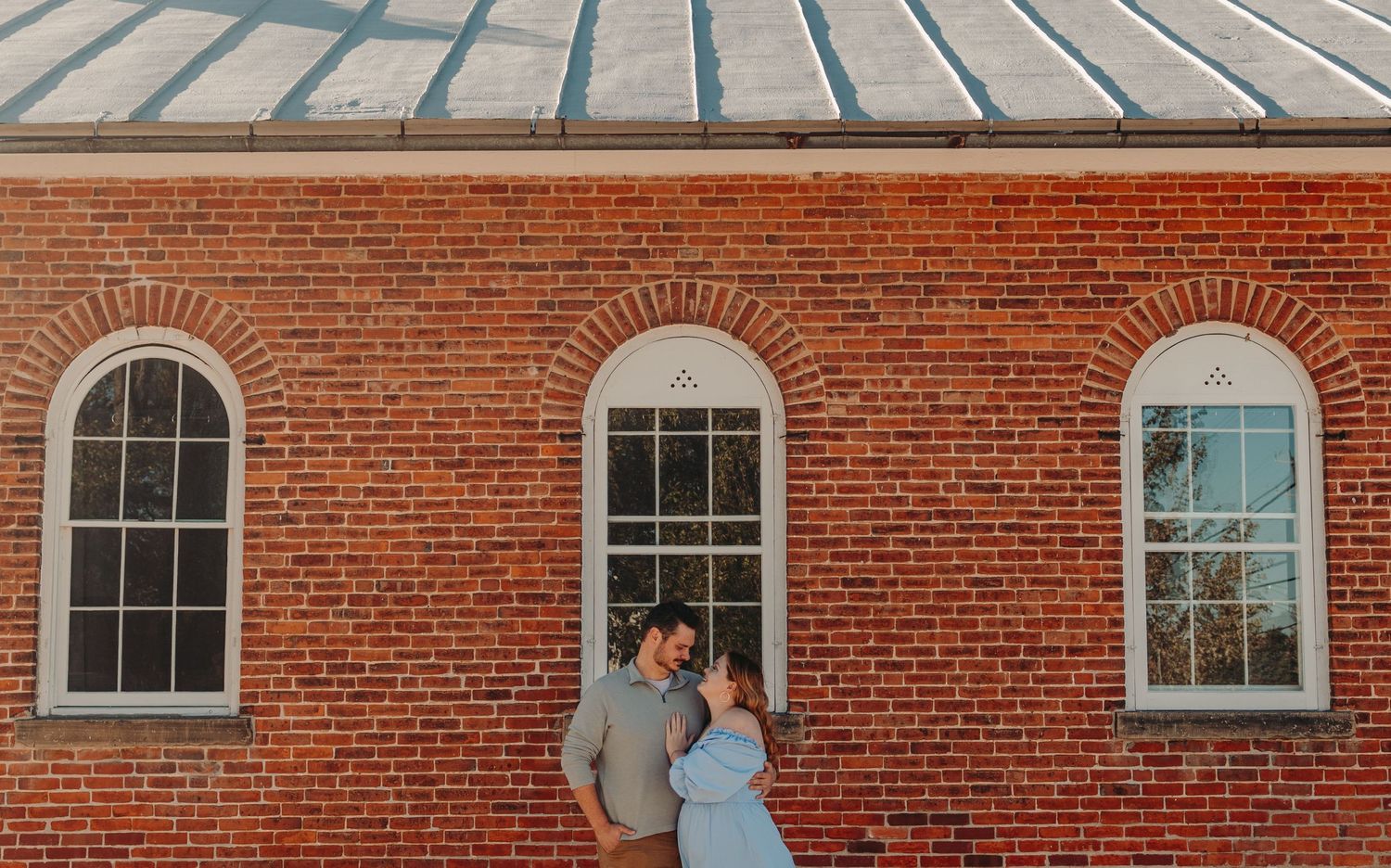 Couple poses romantically against classic red brick wall with arched windows.