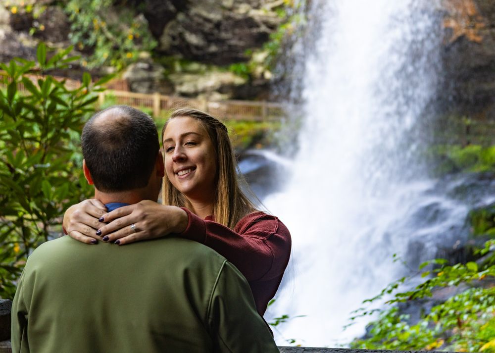 Two people embrace while standing near a cascading waterfall surrounded by lush green foliage.