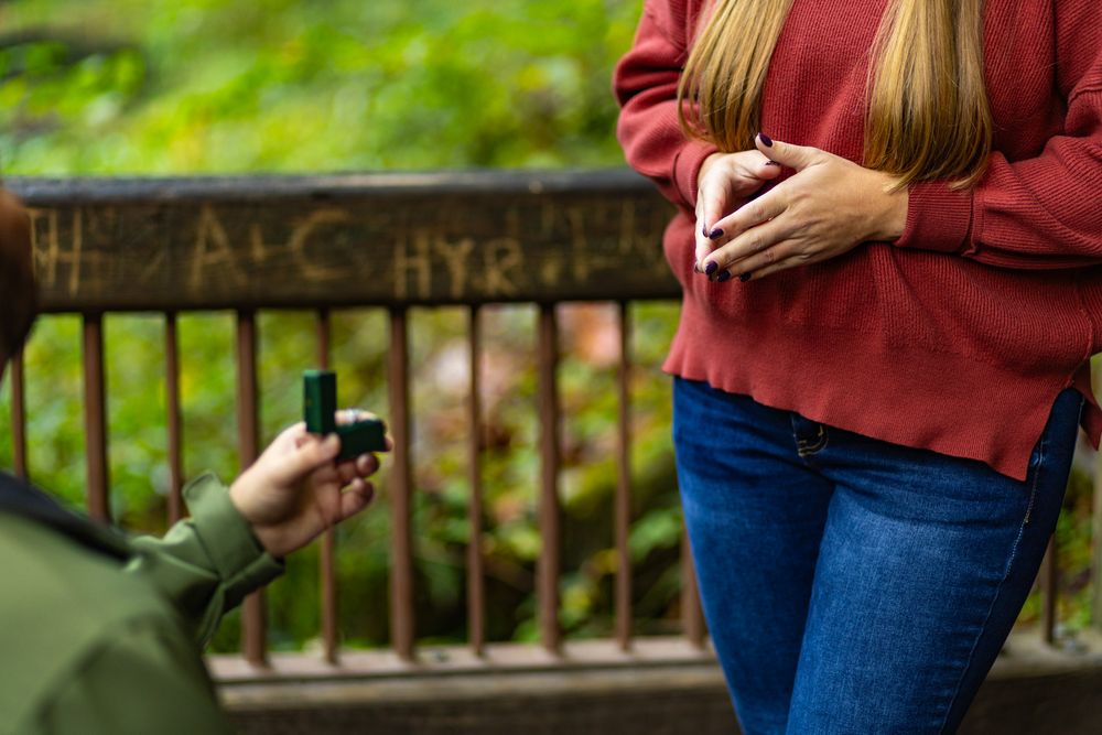 Person in red sweater and jeans standing outdoors with fall foliage in background.