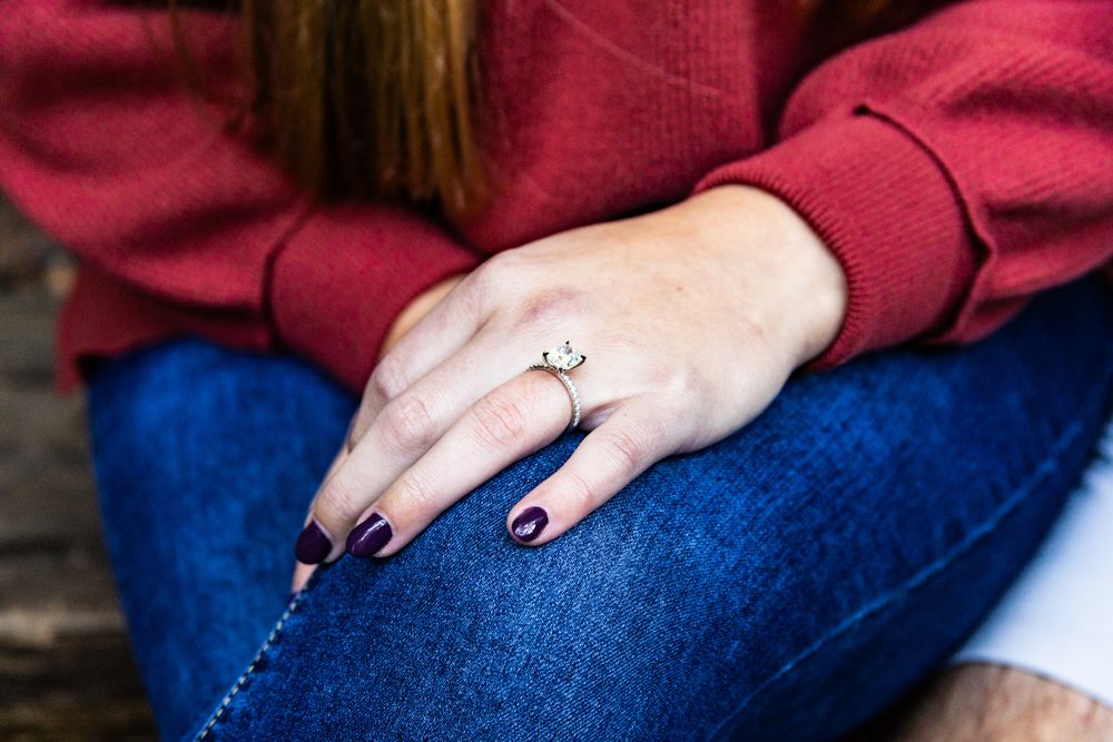 Close-up view of a hand wearing a diamond engagement ring resting on blue jeans with burgundy sweater sleeves visible.