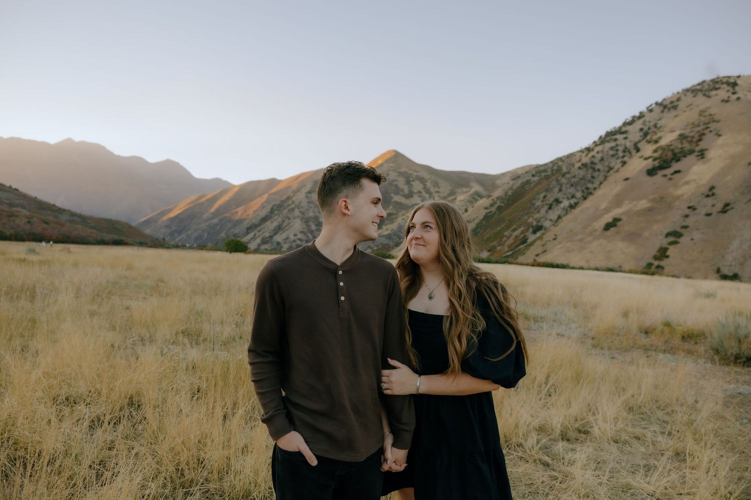 Couple holding hands and smiling in golden grass field with colorful mountain backdrop at sunset.