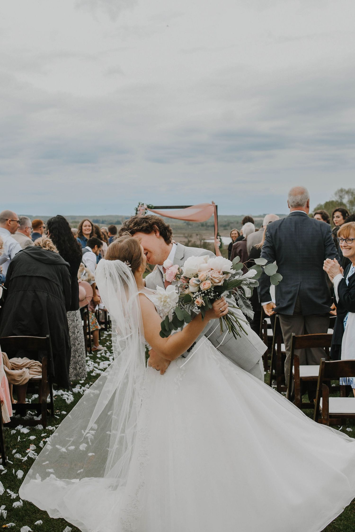 A romantic wedding moment captured outdoors with guests watching as flower petals scatter on the ground during the ceremony.