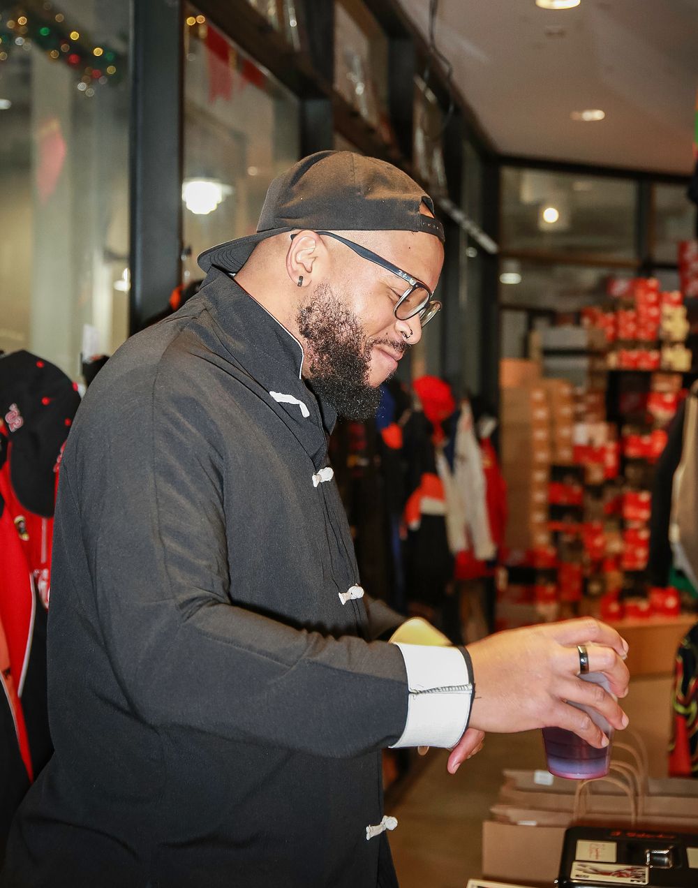 A bearded man in glasses and a black toggle coat stands smiling inside a retail store with holiday decorations.