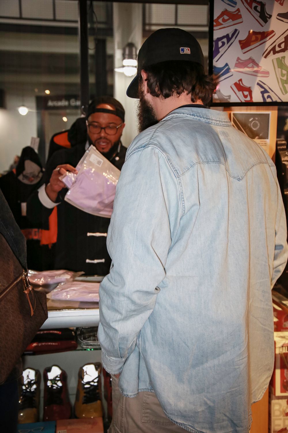 Person in a denim shirt and baseball cap interacts with someone at what appears to be a merchandise counter.