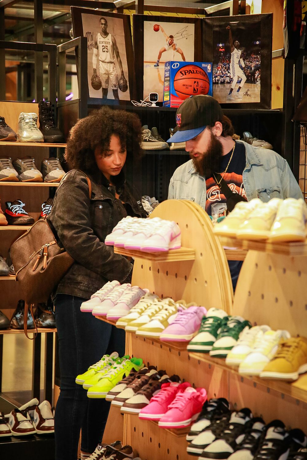 Two customers browse colorful sneakers displayed on wooden shelves at a shoe store with sports memorabilia on the walls.