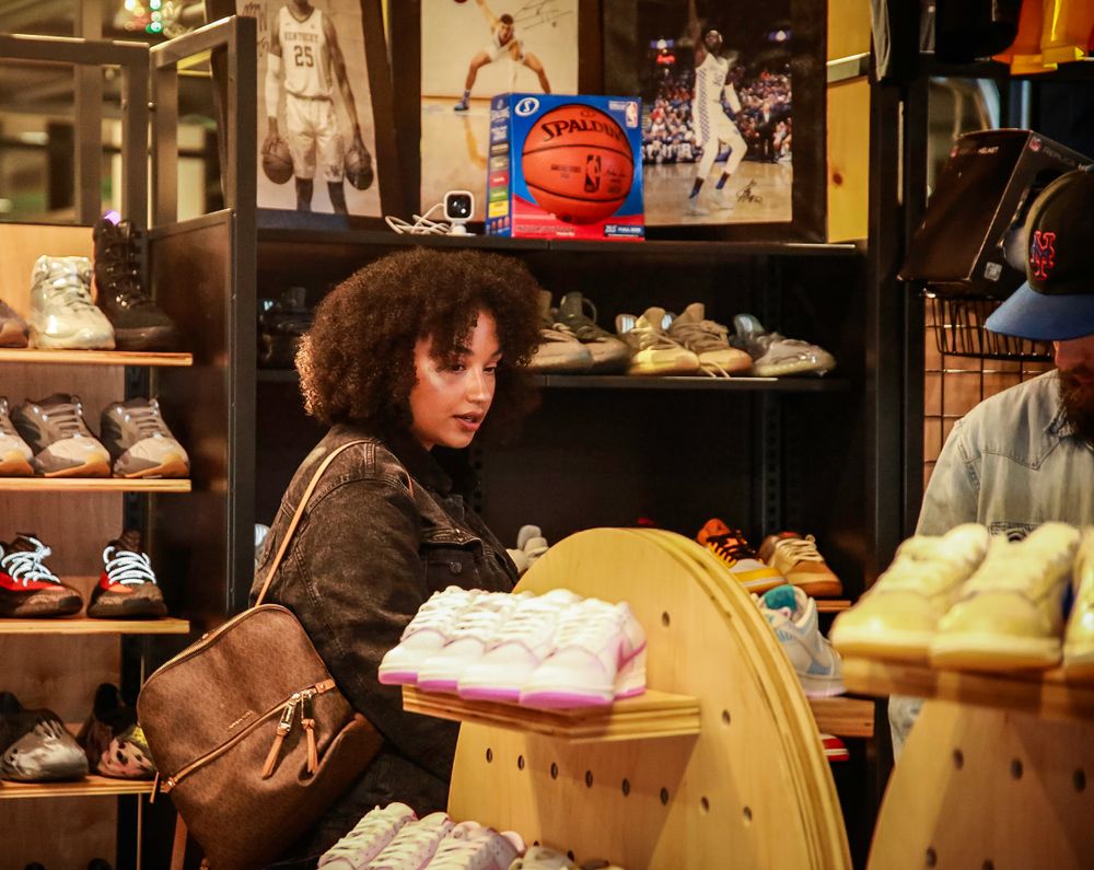 Inside a sneaker store, a person with curly hair browses merchandise while surrounded by shelves of athletic shoes.