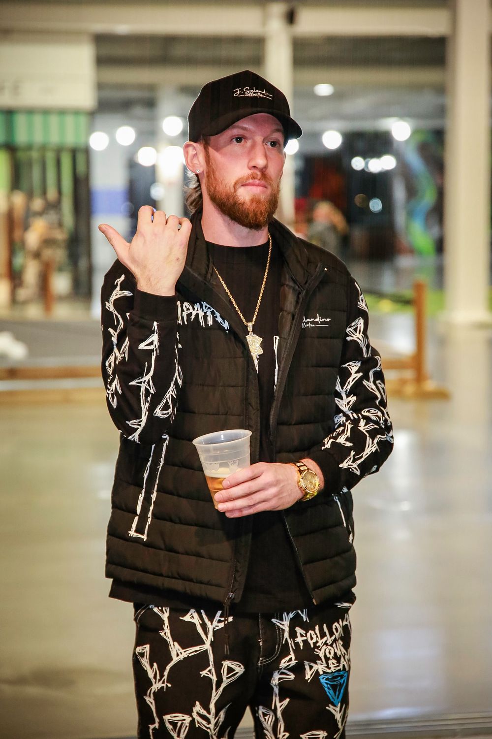 A person wearing a black baseball cap and matching patterned tracksuit holds a drink in an indoor space.