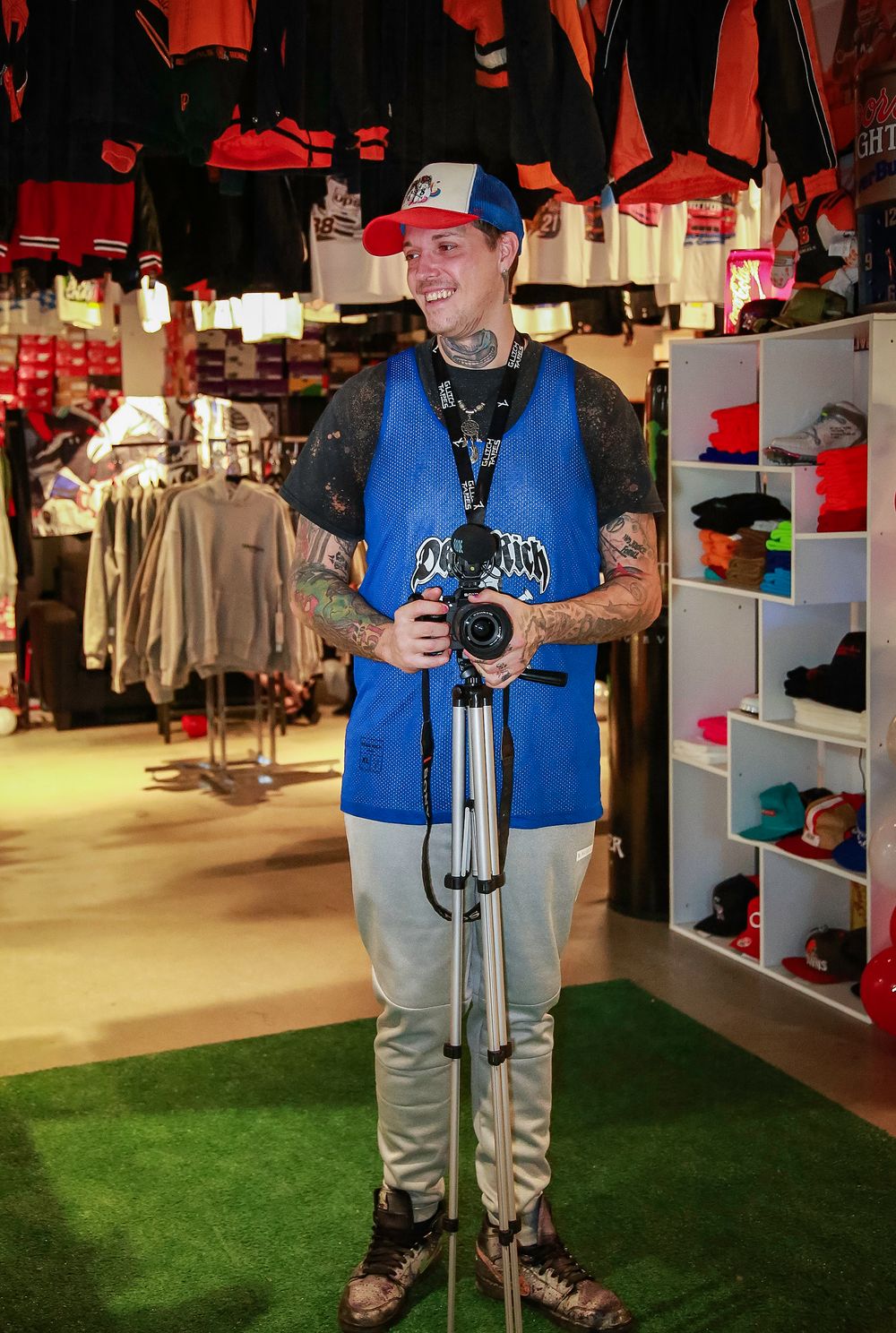 A photographer wearing a blue vest holds a camera on a tripod inside a retail store with red and black merchandise displays.
