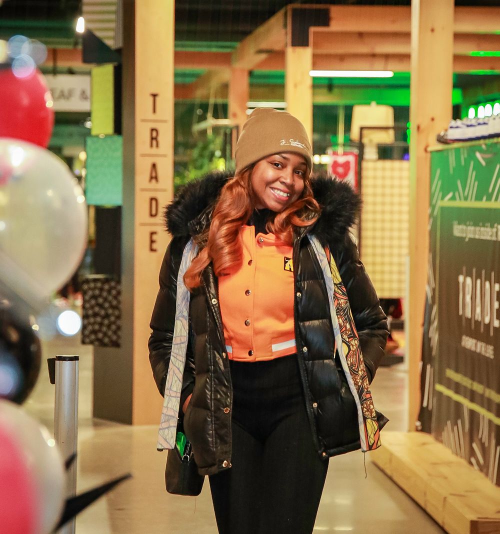 Someone in winter wear including a beanie and fur-trimmed coat walking past store displays and colorful balloons.