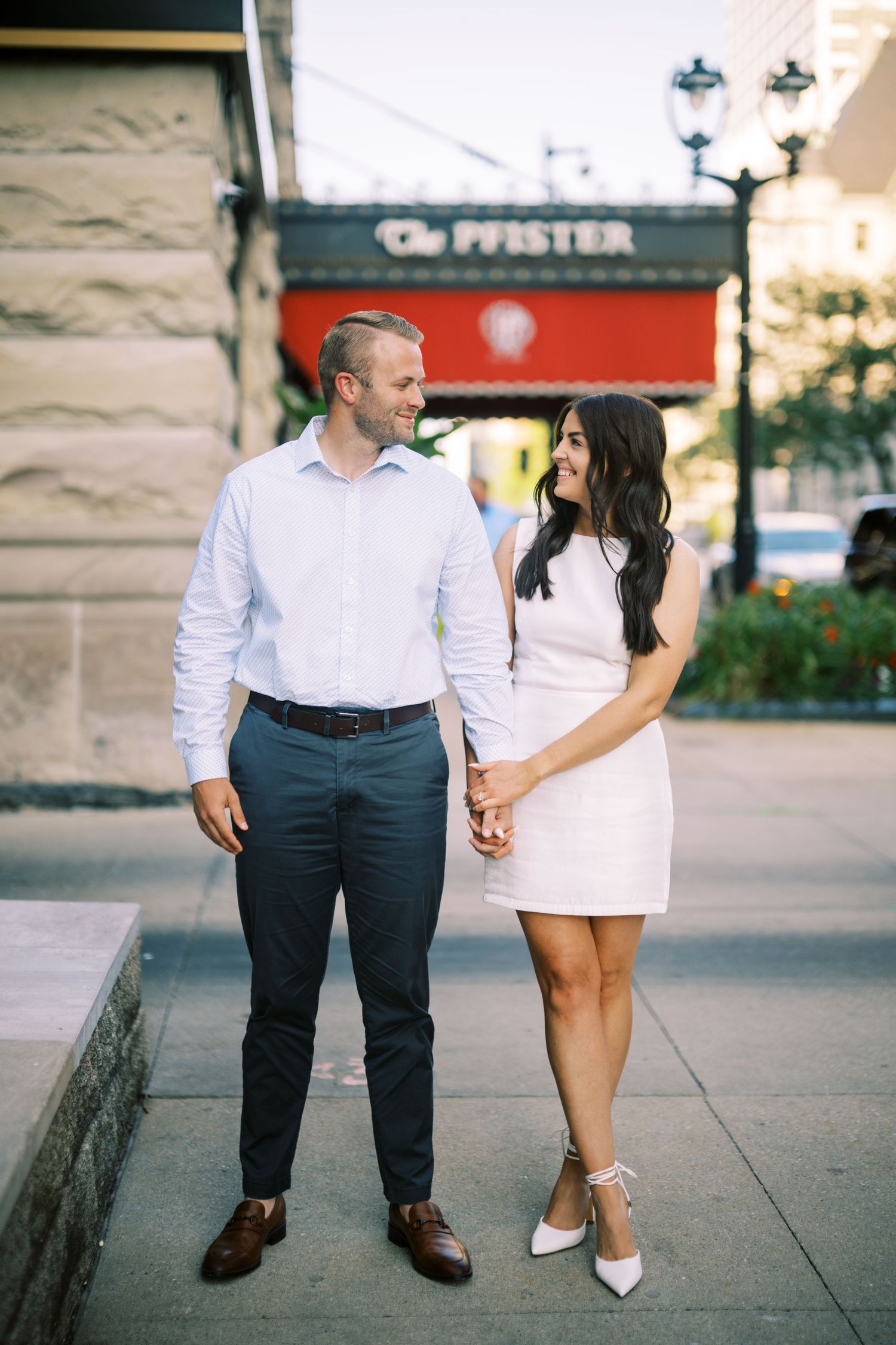 A couple dressed in coordinating white outfits holds hands while standing on a city sidewalk near a storefront.