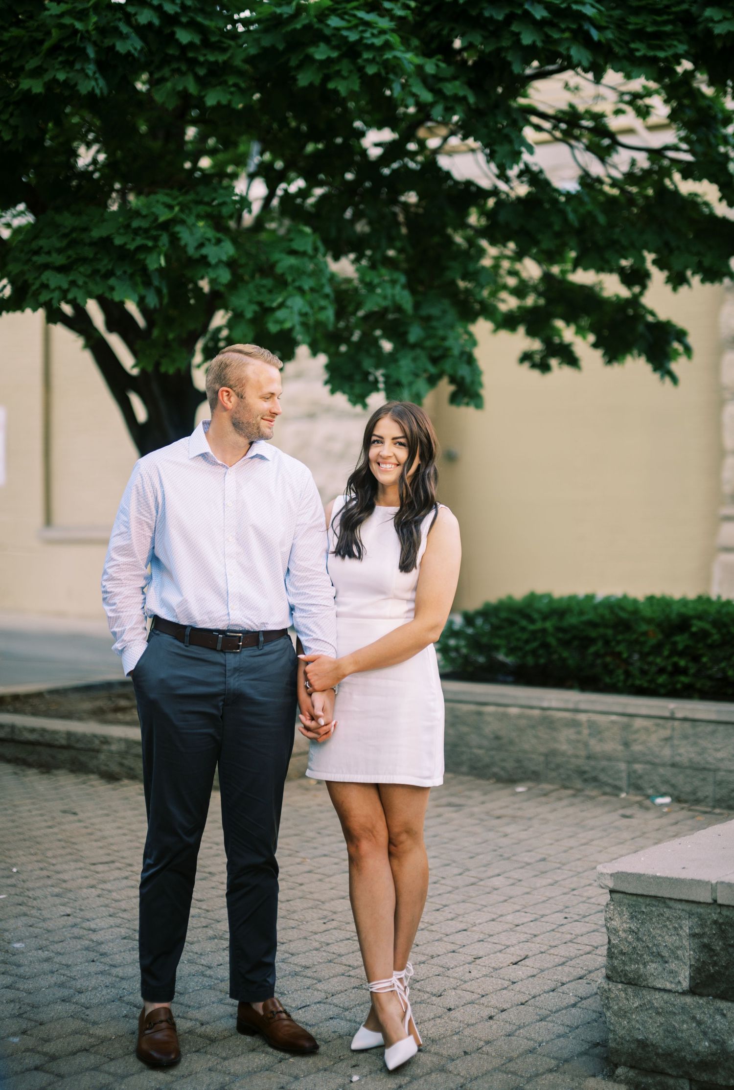 A couple wearing coordinated white and pink outfits poses together on a brick paved walkway under a leafy tree.