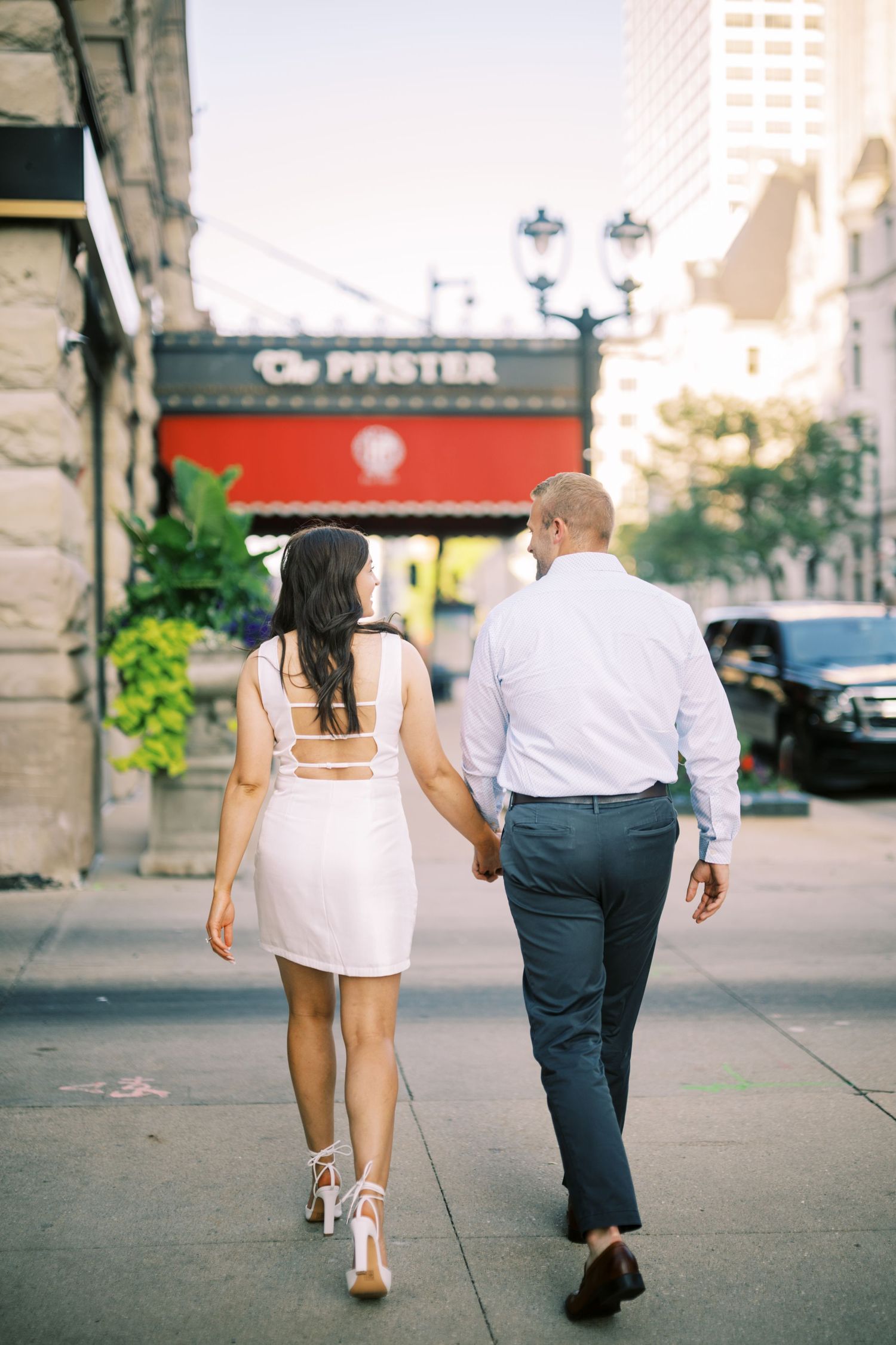 A couple walks hand in hand on a city sidewalk near a restaurant with a red storefront sign.