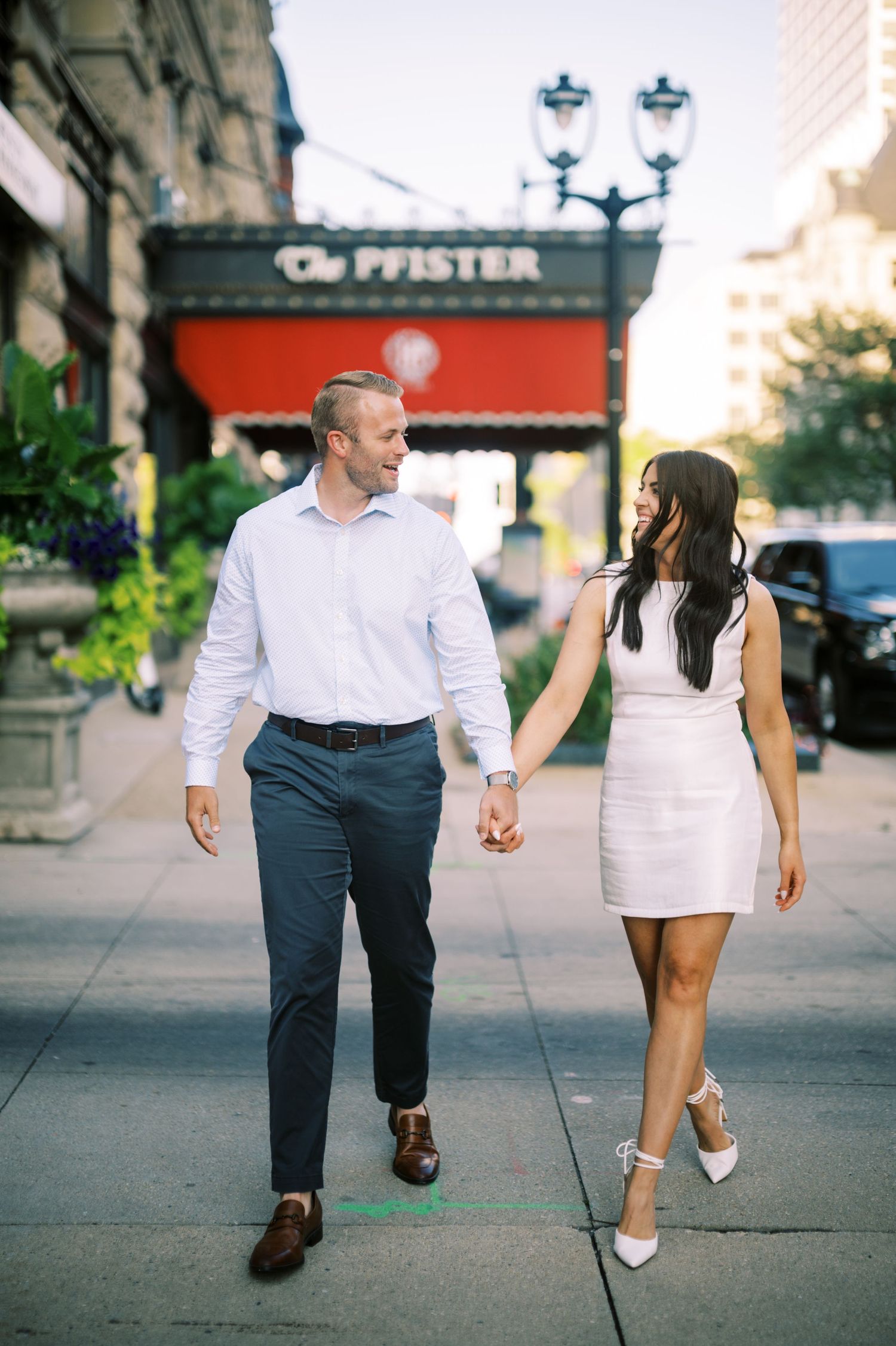 A couple walks hand in hand along a city sidewalk on a sunny day, passing by The Pfister hotel sign.