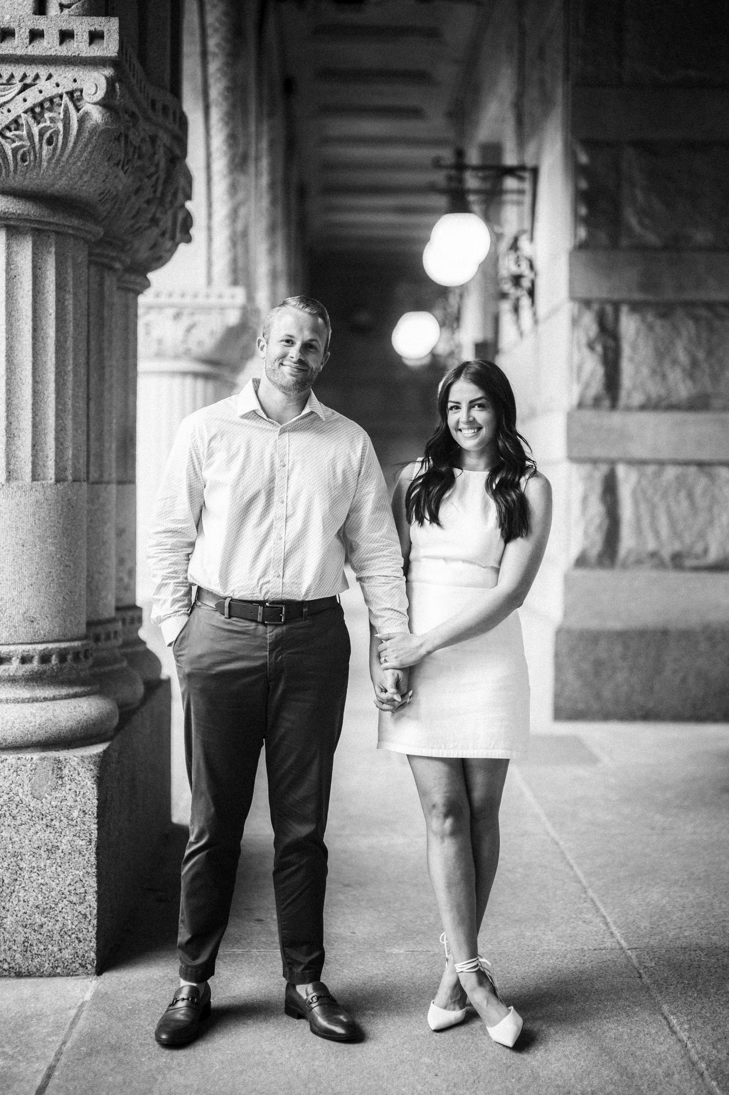 A couple poses in formal attire against ornate stone columns and vintage lighting fixtures in a black and white photo.