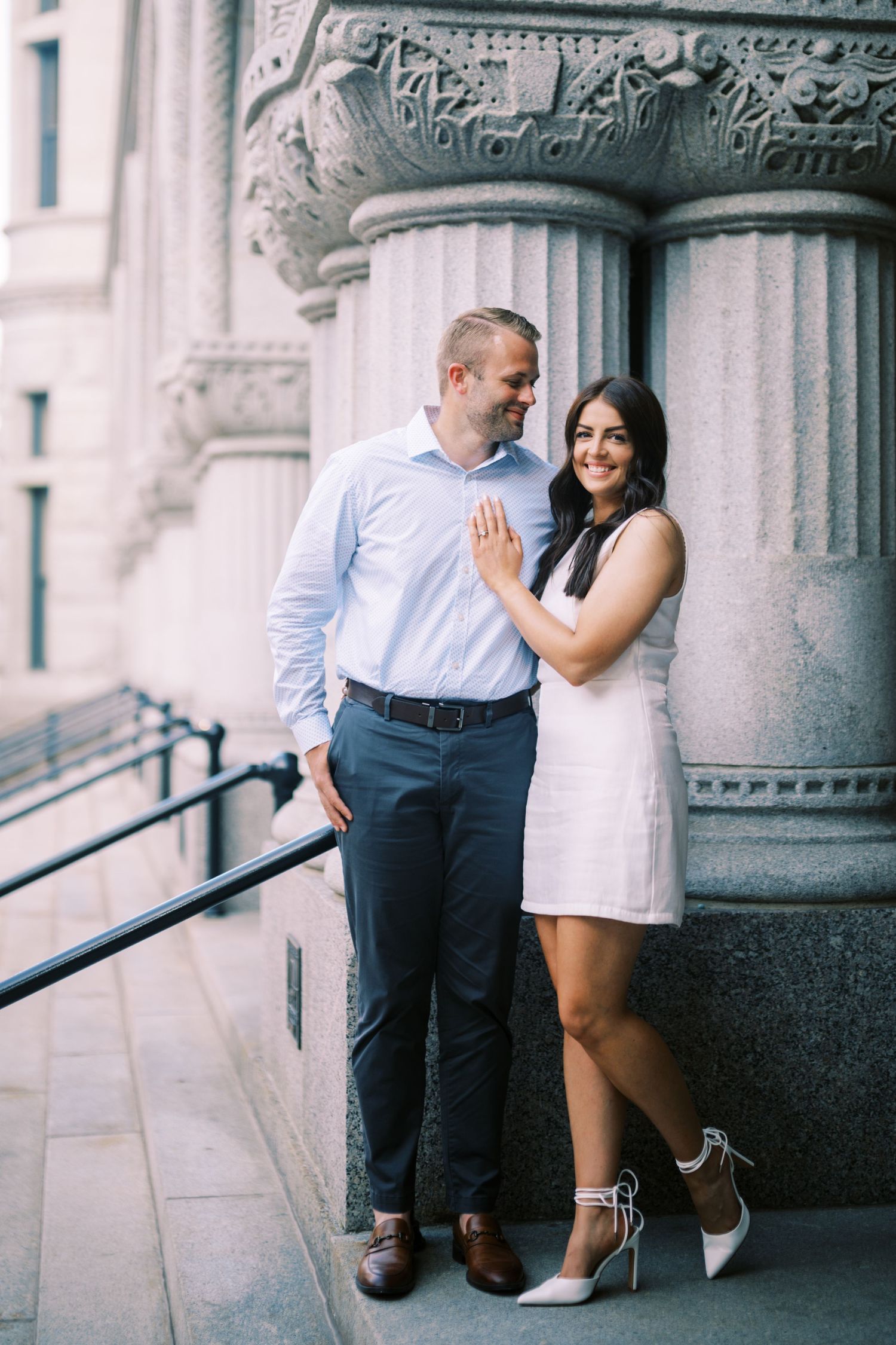 A couple poses together against ornate stone columns on a building exterior in a romantic engagement photo.