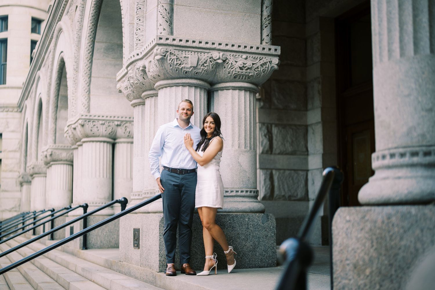 A couple stands together on ornate stone steps of a historic building with elegant columns.