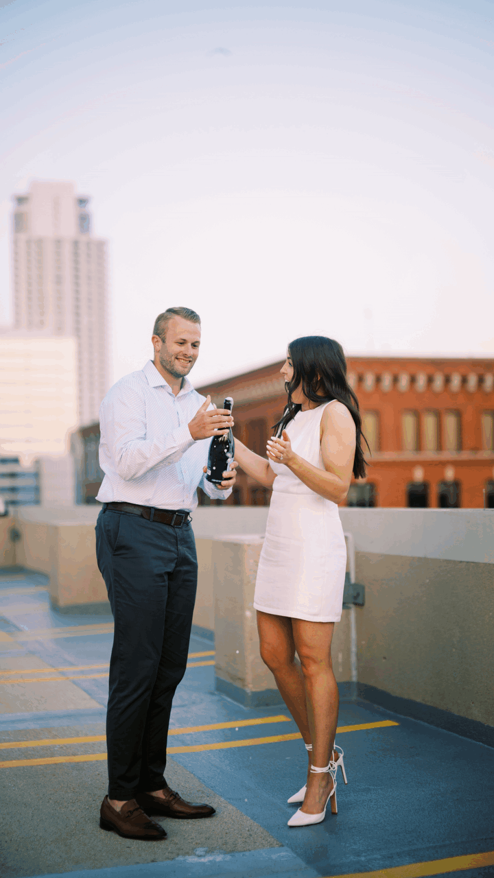 A couple toasts with drinks on a rooftop parking deck with a city skyline view at dusk.