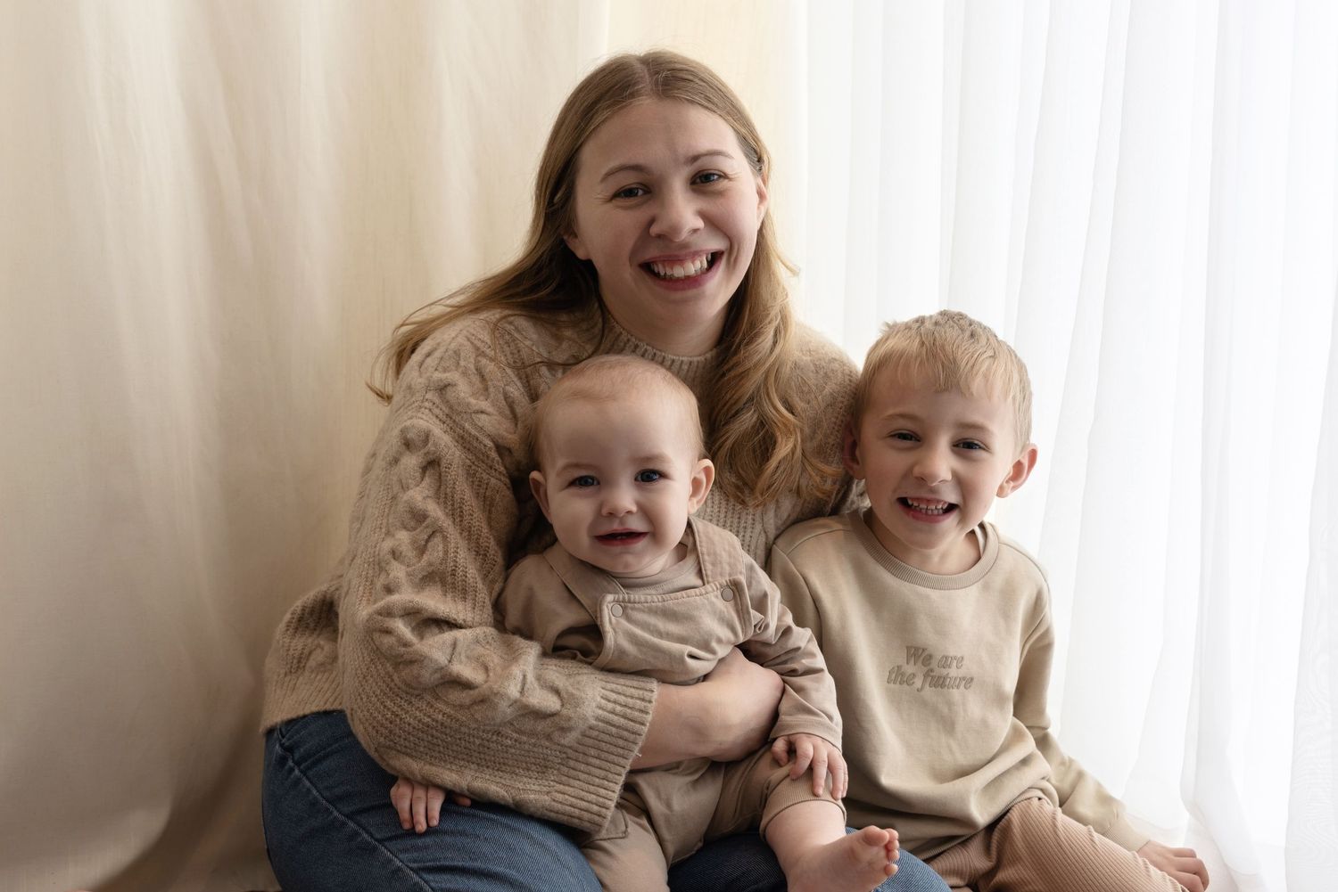 Three people wearing beige sweaters sit together and smile warmly against a light curtain background.