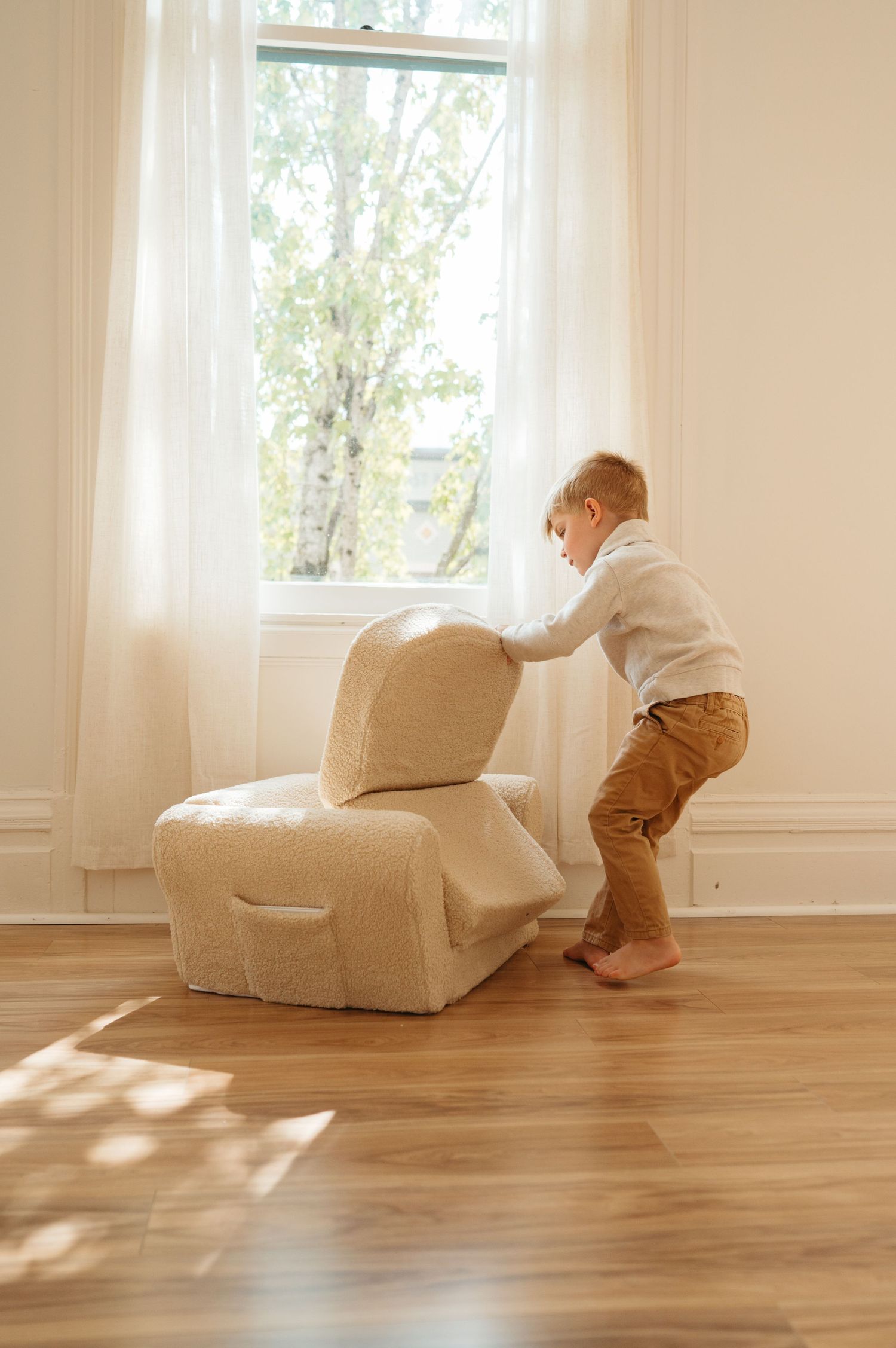 A toddler in brown pants and white top climbs onto a beige armchair in a sunlit room with hardwood floors.