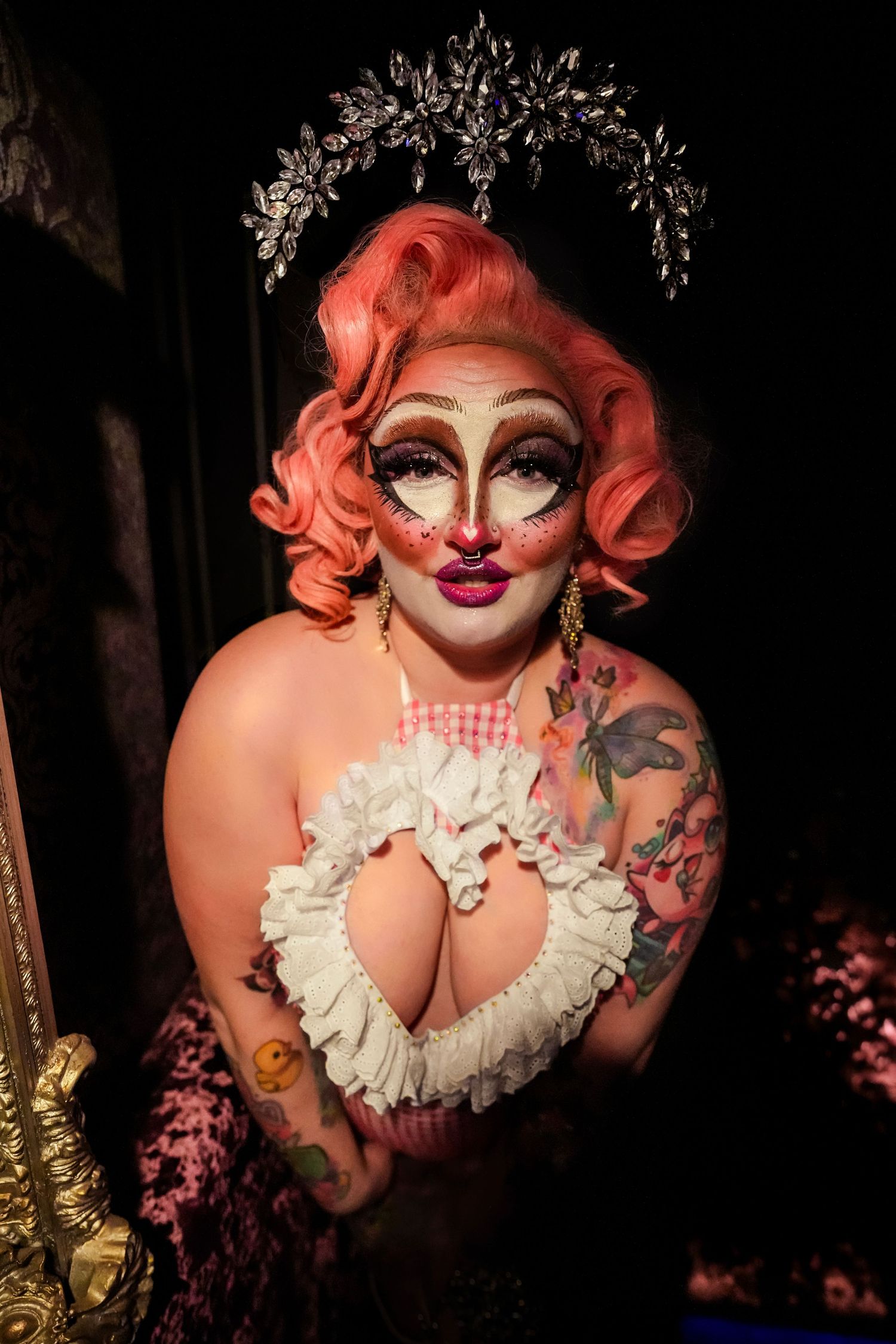 A drag performer in dramatic makeup wearing a white ruffled top poses under decorative floral archway in dim lighting.