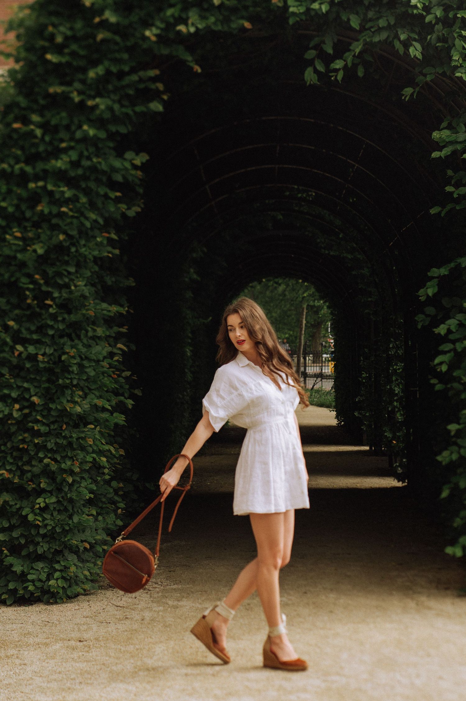 A beautiful woman with red lipstick in a white dress, posing in the Rijksmuseum garden with a green arch in the background.