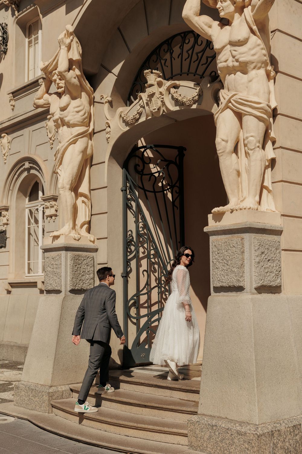 A wedding couple stands near classical stone statues and architecture in warm sunlight.