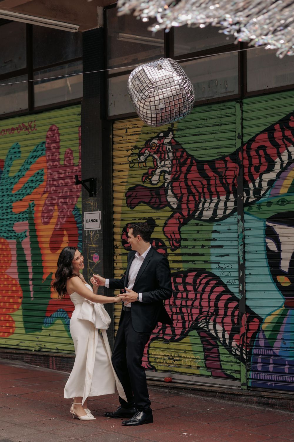 A couple dances under a disco ball against a vibrant street art mural featuring a red tiger.