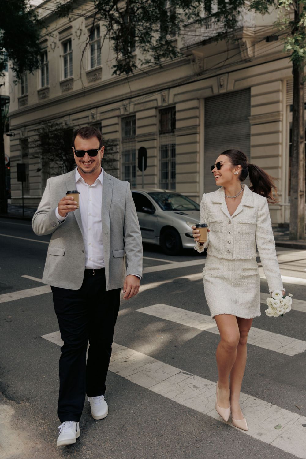 A stylish couple walking across a crosswalk in coordinated outfits while holding coffee cups.