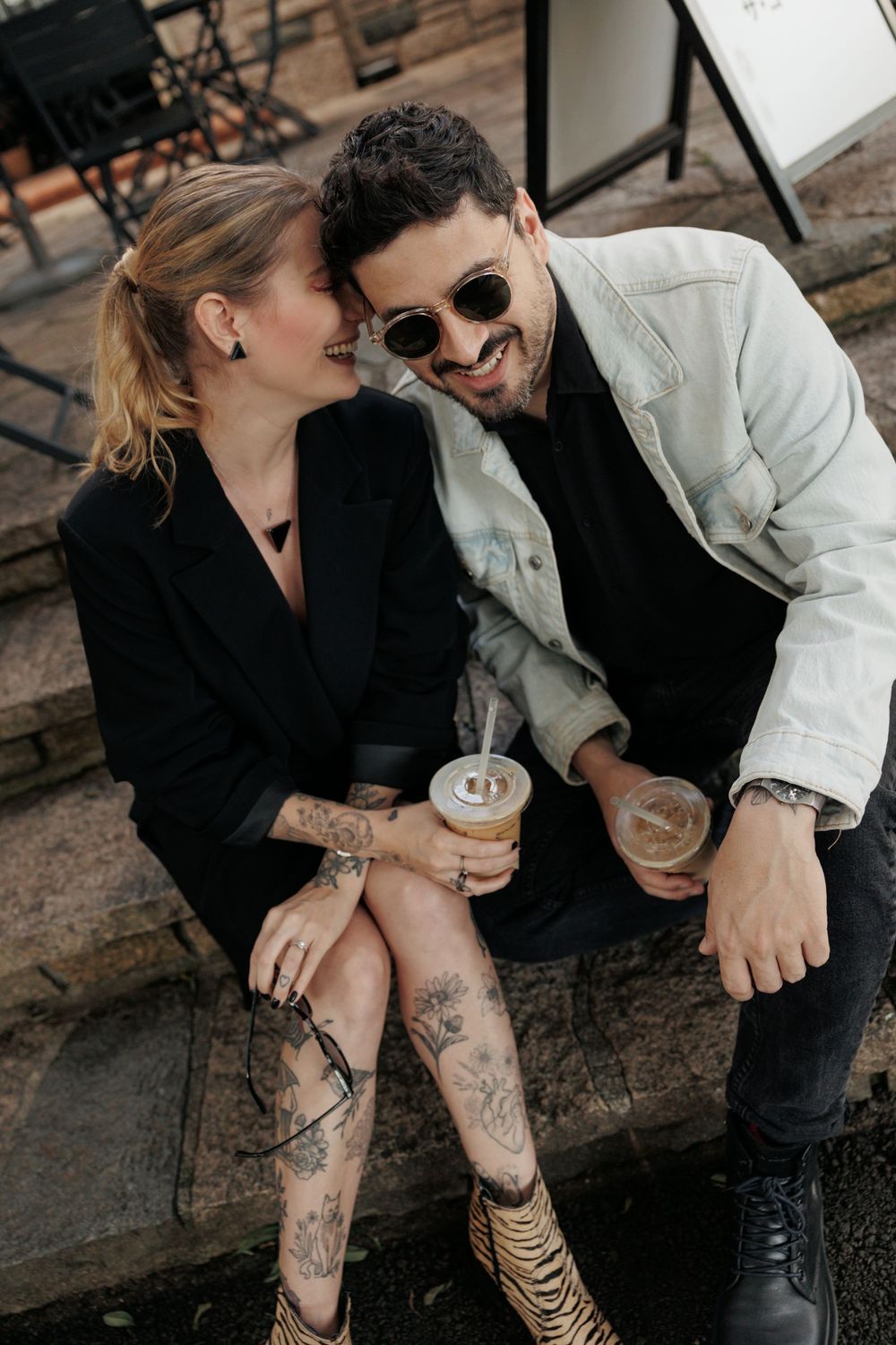 Stylish couple sitting on steps sharing drinks and laughing together, with visible tattoos and fashionable outfits.