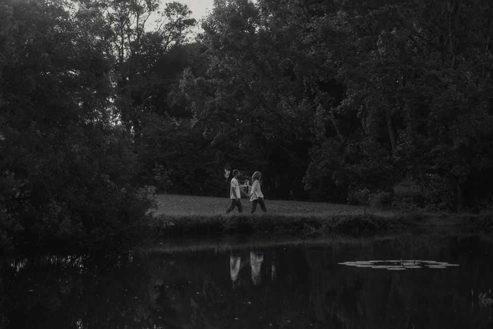 Black and white landscape photo of couple by serene pond with tree reflections.