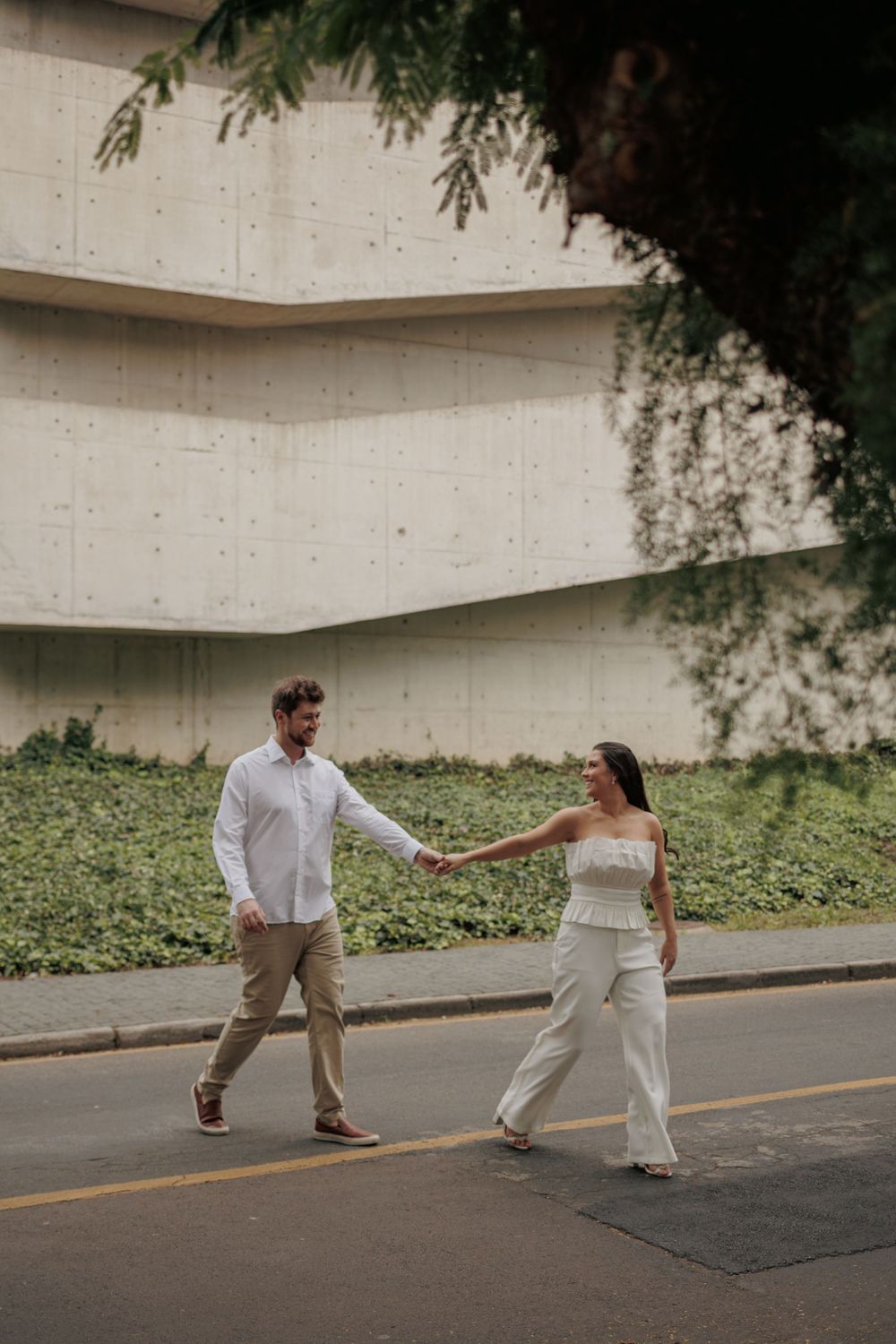 A couple in wedding attire playfully runs and poses on a paved road with trees and a wall in the background.