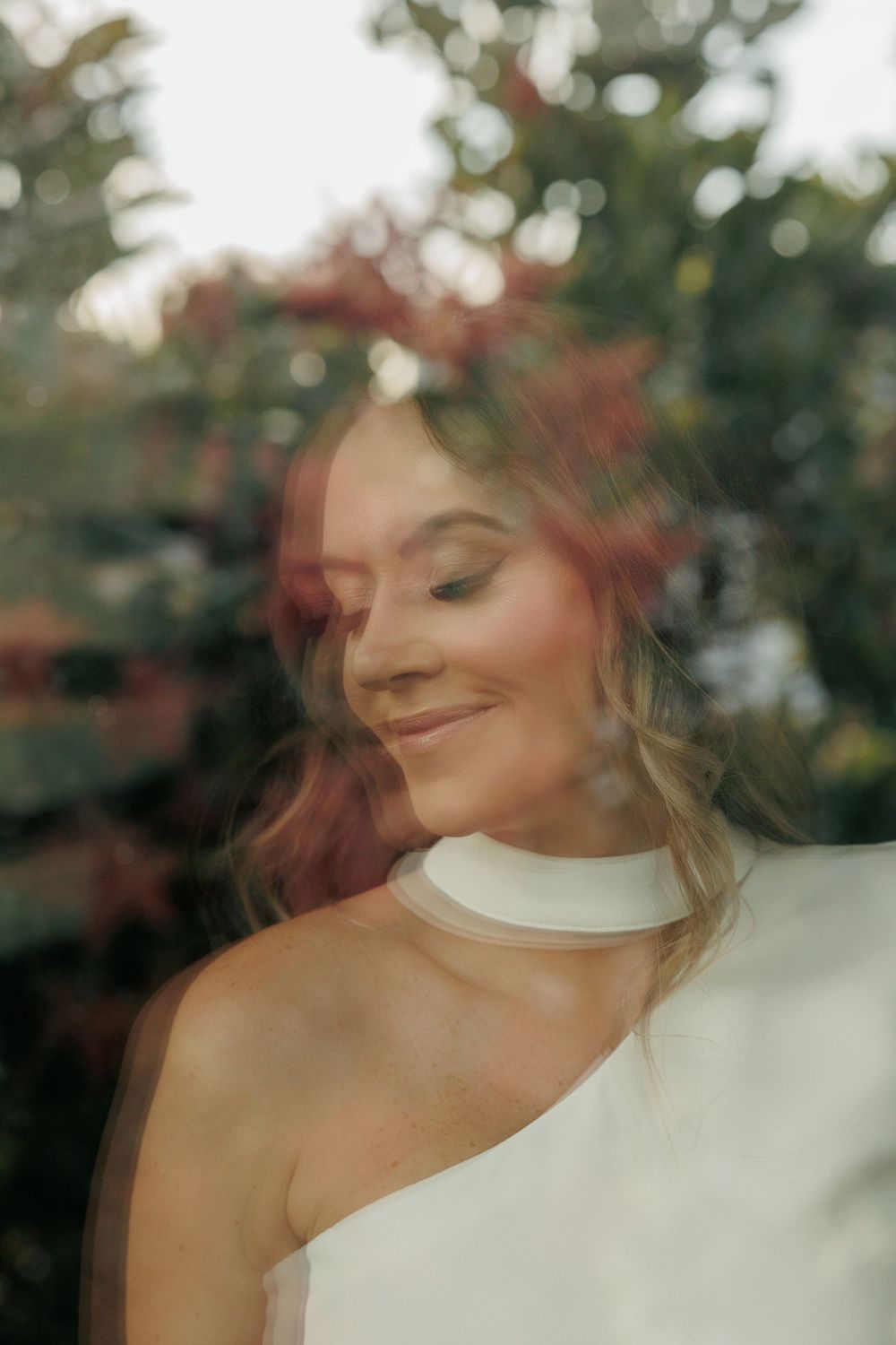 Dreamy portrait through glass with soft natural lighting and foliage backdrop.